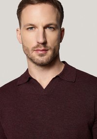 Bordowa dzianinowa koszulka polo z dekoltem w serek i krótkim rękawem. Materiał wydaje się miękki, z subtelną teksturą i czystymi liniami.