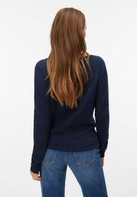 Top a maniche lunghe di un blu navy testurizzato con una silhouette aderente, abbinato a jeans azzurri. La vista posteriore mostra il tessuto liscio e l'orlo arrotondato.
