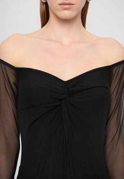 Melna off-shoulder blūze ar savītu mezglu centrā, kurai ir caurspīdīgas garās piedurknes un gluda, pieguloša korpusa daļa.
