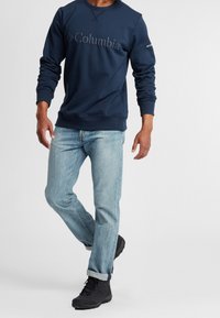 Sweatshirt navy con logo del marchio testurizzato, abbinato a jeans azzurri chiari e stivali neri impermeabili, con orli arrotolati e vestibilità rilassata.