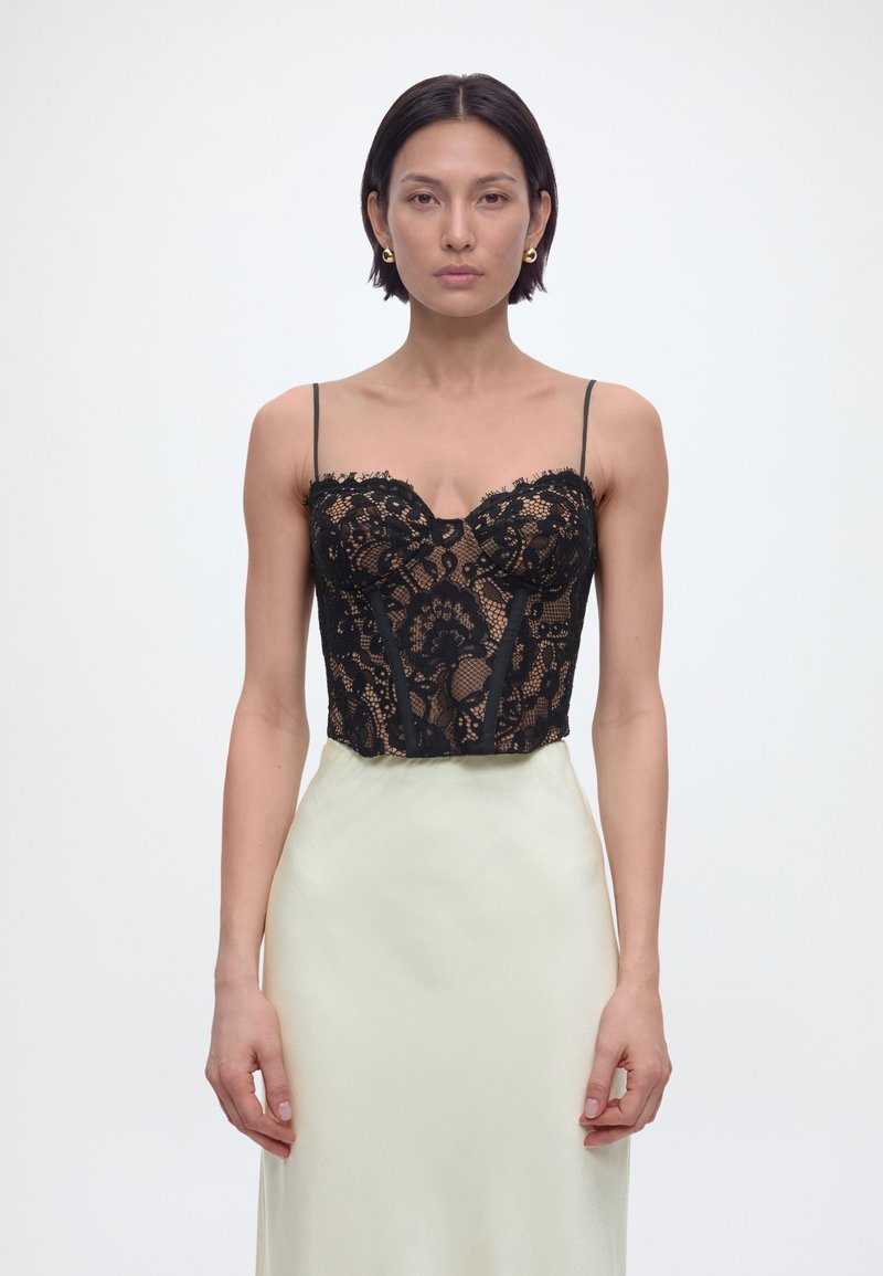 Γυναίκα με κοντά σκούρα μαλλιά, φορώντας μαύρο κορσέ από δαντέλα και σατέν φούστα ανοιχτού χρώματος, που στέκεται μπροστά σε ένα απλό λευκό φόντο.