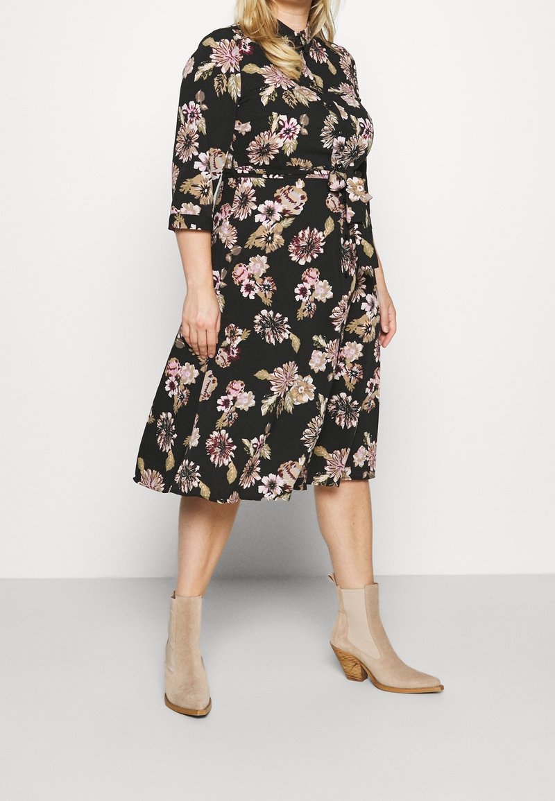 Black floral dress with three-quarter sleeves, cinched waist, knee-length skirt, featuring pink and cream flowers. Beige ankle boots with a block heel.