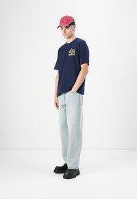 Young man standing, wearing red cap, navy graphic t-shirt, light blue jeans, and black chunky shoes against plain white background.
