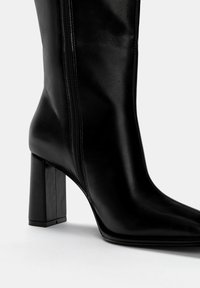 Schwarzer Lederknöchelschuh mit spitzer Zehe, Blockabsatz und seitlichem Reißverschluss. Glatte Textur mit minimalistischen Designelementen.