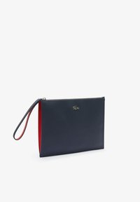 Navy clutch torbica z gladko teksturo, z rdečim notranjim delom, z zadrgo in ročajem za zapestje. Vidna je majhna kovinska nalepka.