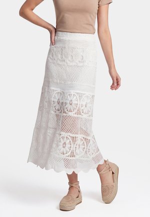Frau trägt einen weißen Spitzen-Midirock mit Blumenmustern, ein beiges kurzärmliges Oberteil und beige Espadrilles mit Knöchelriemen.