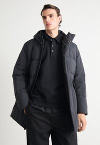 Black puffer jacket with a hood, featuring quilted texture, zip closure, and snap buttons. Worn over a black polo shirt and pants.