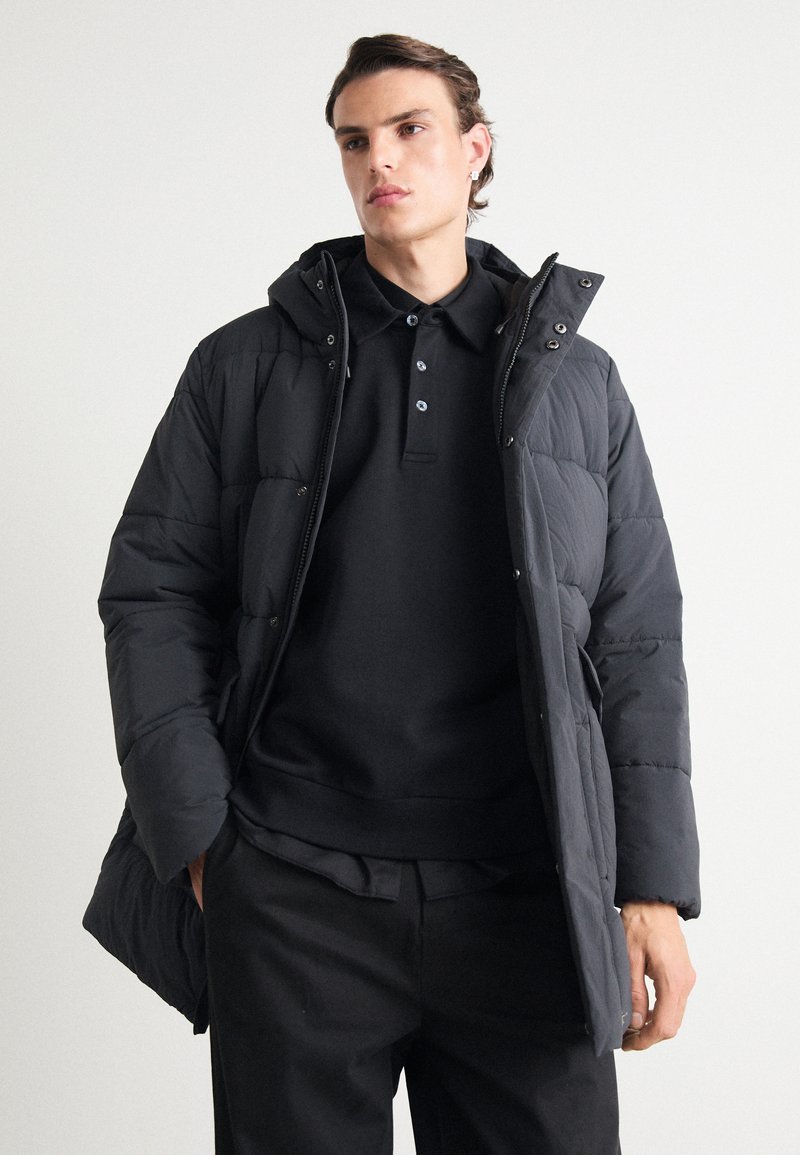 Black puffer jacket with a hood, featuring quilted texture, zip closure, and snap buttons. Worn over a black polo shirt and pants.