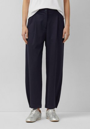 Kobieta w luźnych granatowych spodniach i srebrnych sneakersach, stojąca na tle jednolitego jasnoszarego tła.