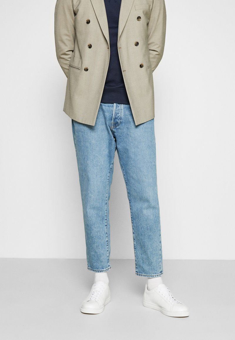 Beige Doppelreiher-Blazer über einem marineblauen Pullover, kombiniert mit hellblauen gerade geschnittenen Jeans und weißen Sneakers. Enthält strukturierter Stoff und sichtbare Nähte.