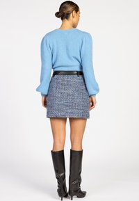 Vrouw in een lichtblauwe fluffy trui, blauwe tweed minirok met een zwarte riem en kniehoge zwarte leren laarzen, staand met haar gezicht naar weg.