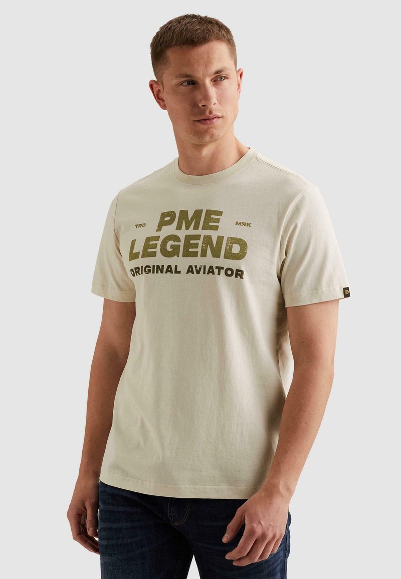 Νέος άνδρας με μπεζ μπλουζάκι PME Legend Original Aviator και σκούρο τζιν, κοιτάζει ελαφρώς πλάγια σε απλό φόντο.