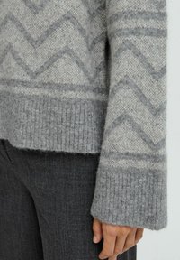 Gros plan sur une main reposant sur le côté, portant un pull en tricot gris à motifs et un pantalon gris foncé.