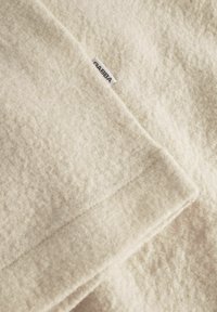 Krémová vlněná deka s měkkým, texturovaným povrchem. Obsahuje malou textilní etiketu se značkou "GABBA."