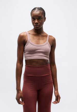 Femme aux cheveux tressés portant un haut court mauve et un legging taille haute marron foncé, debout devant un fond gris uni.