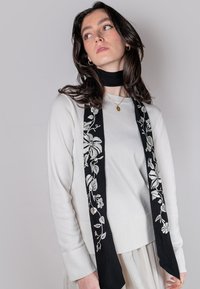 Weißer Pullover mit langem schwarzem Schal, der auf einer Seite weiße florale Stickereien aufweist. Das Modell präsentiert einen neutralen Hintergrund mit sanfter Beleuchtung.