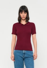 Donna con capelli scuri alla spalla, che indossa una polo corto a maniche corte rigata color bordeaux e jeans blu, in piedi davanti a uno sfondo bianco.