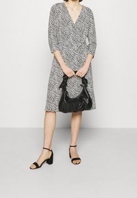 Schwarz-weißer gepunkteter Wickelkleid mit Drei-Viertel-Ärmeln und tailliertem Schnitt. Hält eine schwarze zusammengeknüllte Handtasche und trägt schwarze Riemchensandalen.
