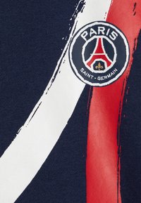 Granatowy materiał z okrągłym logo z napisem "PARIS" i "SAINT-GERMAIN", uzupełniony białymi i czerwonymi akcentami w stylu pędzla.