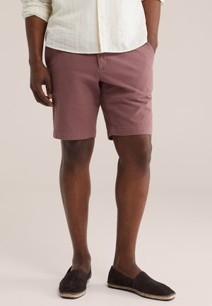 Férfi poros rózsaszín rövidnadrágban, krémszínű hosszú ujjú ingben és fekete bebújós espadrille cipőben, zsebében tartva a kezét, áll.