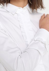 White shirt made of lightweight fabric featuring a ruffled placket and decorative cuffs. The collar is classic with button closures.