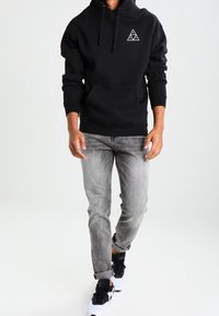 Czarna bluza z kapturem z kieszenią z przodu i białym logo w kształcie trójkąta, zestawiona z jasnoszarymi dżinsami i czarno-białymi trampkami.