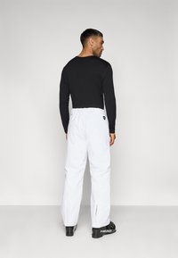 Λευκό παντελόνι χιονιού με ρυθμιζόμενη μέση, σε συνδυασμό με ένα μαύρο μακρυμάνικο πουκάμισο. Το μοντέλο κρατάει μια μαύρη μπότα σκι.