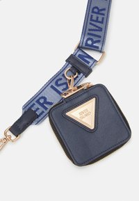 Čtvercová peněženka na mince v námořnické modři se zlatým zipem a připojenou trojúhelníkovou zlatou nášivkou loga na modrém popruhu s logem.