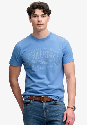 Ung mand iført en blå Superdry t-shirt, blå jeans, brun læderbælte og armbåndsur på en ensfarvet hvid baggrund.