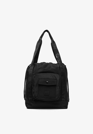 Czarna torba typu tote z szerokimi paskami, przednia kieszeń na suwak z klapką zapinaną na guzik oraz prostokątnym spodem, wykonana z miękkiej tkaniny.