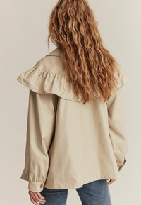 Beige langærmet jakke med flæsekant og knaplukninger ved ærmerne, lavet af glat stof. Har en afslappet pasform og falder under taljen.