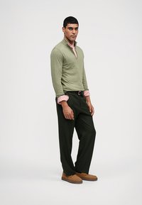 Grön zip-top över en rosa kragskjorta, kombinerat med mörkgröna byxor och bruna skor. Slät textur och avslappnad design.