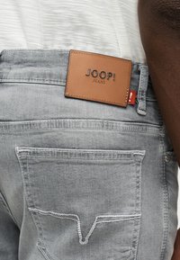 Πίσω όψη ατόμου που φοράει ανοιχτό γκρι τζιν με ταμπέλα από ταμπά δέρμα με την ένδειξη «JOOP! JEANS» και κόκκινη ετικέτα με θαυμαστικό.