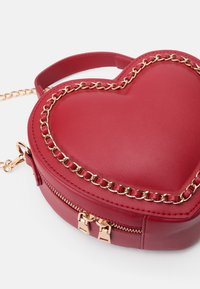 Sac en forme de cœur rouge en simili cuir lisse. Caractéristiques d'accents en chaîne dorée et d'une fermeture à double zip sur le côté.