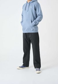 Felpa con cappuccio di colore azzurro con tasca a marsupio e piccolo logo sul petto, abbinata a pantaloni neri a vestibilità rilassata e sneakers blu.