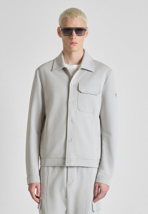 Jeune homme aux cheveux blonds courts portant de grandes lunettes de soleil noires, une veste gris clair boutonnée avec une poche poitrine, et un pantalon assorti.