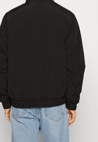 Černá prošívaná bunda s žebrovaným lemem a manžetami. Má hladký, matný povrch. Nošena přes světle modré džíny.