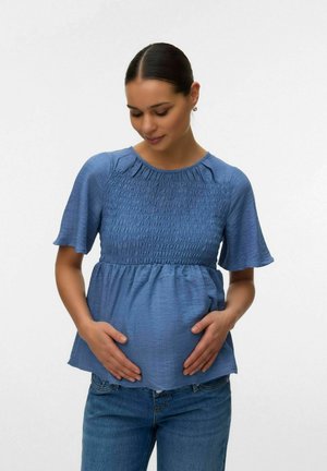 Femme enceinte portant un chemisier bleu texturé et un jean, tenant doucement son ventre, regardant vers le bas avec un doux sourire sur un fond uni.