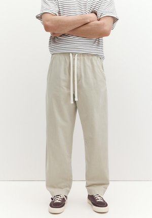 Persona in piedi con le braccia incrociate, indossa pantaloni beige con coulisse, una maglietta a maniche corte a righe e scarpe da ginnastica scure su uno sfondo bianco uniforme.