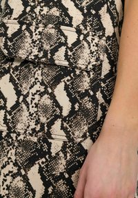 Beige stof met zwart slangprint, met een textuurpatroon. Een klein zakje is zichtbaar, geaccentueerd door witte stiksels.
