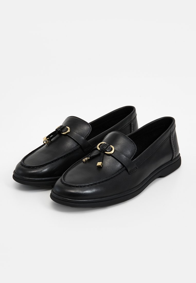 Mocassins en cuir noir avec boucle dorée et détail à pampilles, présentant un bout arrondi et une semelle plate basse, présentés sur fond blanc.