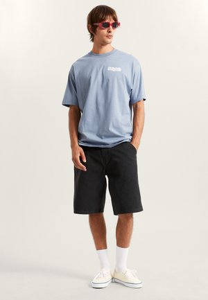 T-shirt en coton bleu clair avec un logo, assorti à un short en denim noir, des chaussettes blanches et des baskets légères. Le modèle porte des lunettes de soleil rouges.