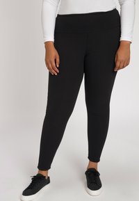 Zwarte leggings met een hoge tailleband, gemaakt van een zachte, rekbare stof. Het model draagt zwarte sneakers met witte zolen.