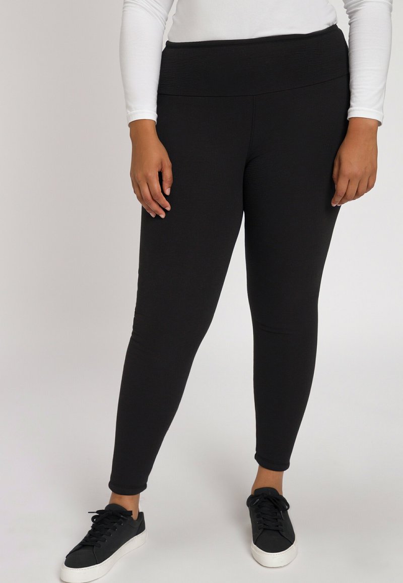 Zwarte leggings met een hoge tailleband, gemaakt van een zachte, rekbare stof. Het model draagt zwarte sneakers met witte zolen.