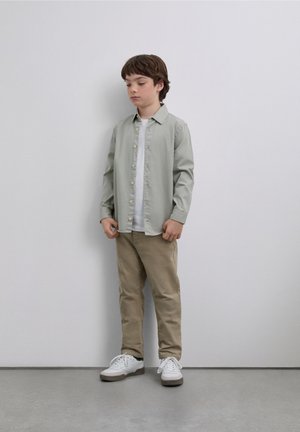 Camisa de botones verde claro, camiseta blanca debajo, pantalones beige y zapatillas blancas con detalles grises. De pie sobre un suelo gris liso.