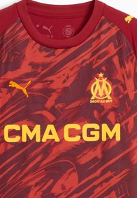 Röd och mörkröd kortärmad tröja med en strukturerad design. Har gula logotyper: "Puma," "CMA CGM," och en klubbemblem med stjärnor.