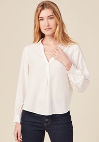 Weiße Bluse mit langen Ärmeln, Kragen, Knopfleiste und strukturiertem Stoff; kombiniert mit dunkelblauen Jeans in enger Passform.