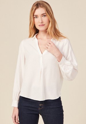 Weiße Bluse mit langen Ärmeln, Kragen, Knopfleiste und strukturiertem Stoff; kombiniert mit dunkelblauen Jeans in enger Passform.