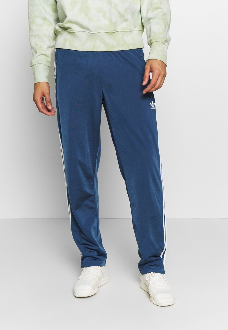 Modré sportovní kalhoty s bílými bočními pruhy, vyrobené z hladkého materiálu, doplněné zelenou mikinou s tie-dye vzorem a bílými teniskami.