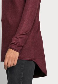 Burgunderfarbenes Langarmshirt mit abgerundetem Saum, strukturiertem Stoff und taillierten Ärmeln, kombiniert mit dunklen Jeans.